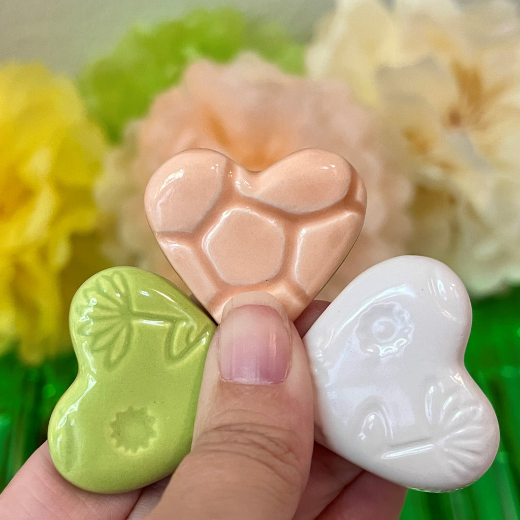 Three heart-shaped ceramics in green, pink, and white held by a hand with a blurred floral background.