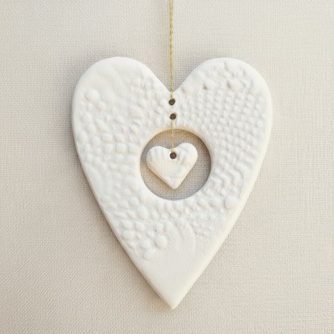 A white ceramic heart-shaped ornament with a smaller heart suspended in the center, displayed against a fabric background.