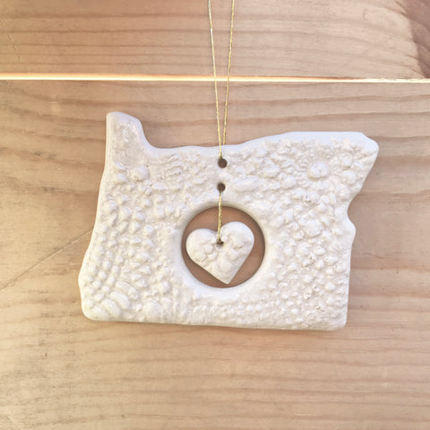 A white ceramic ornament in the shape of Oregon with a heart in the center, hanging from a string.