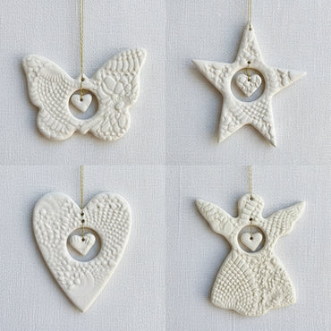 Four white ceramic ornaments shaped like a butterfly, star, heart, and angel on a fabric surface.