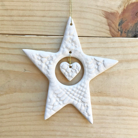 A white ceramic star with a heart-shaped cutout in the center, hanging on a string against a wooden background.