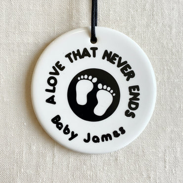 A round, white ceramic footprint ornament with the words 'A LOVE THAT NEVER ENDS' and a space for a name.