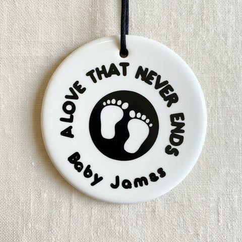 A round, white ceramic footprint ornament with the words 'A LOVE THAT NEVER ENDS' and a space for a name.