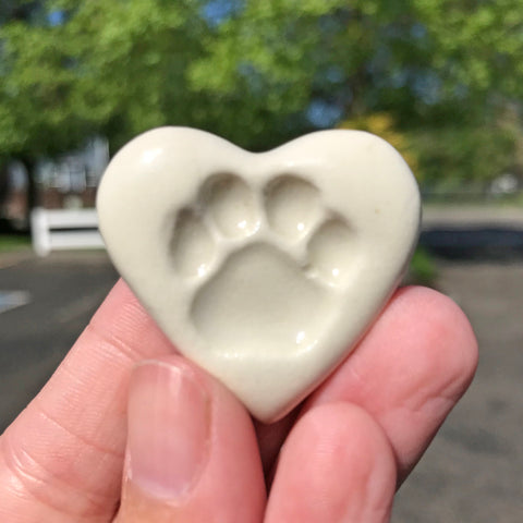 A hand holding a beige ceramic heart-shaped ceramic with a paw print design.