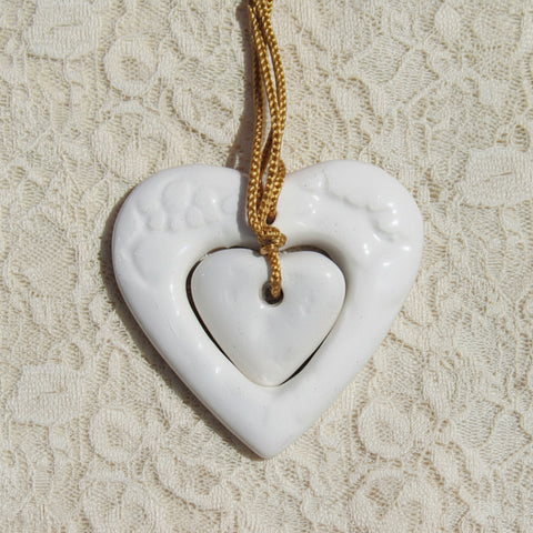 Two handcrafted ceramic hearts in white color, with one smaller heart nested inside the larger one, attached to a gold-colored silk cord, set against a lace background